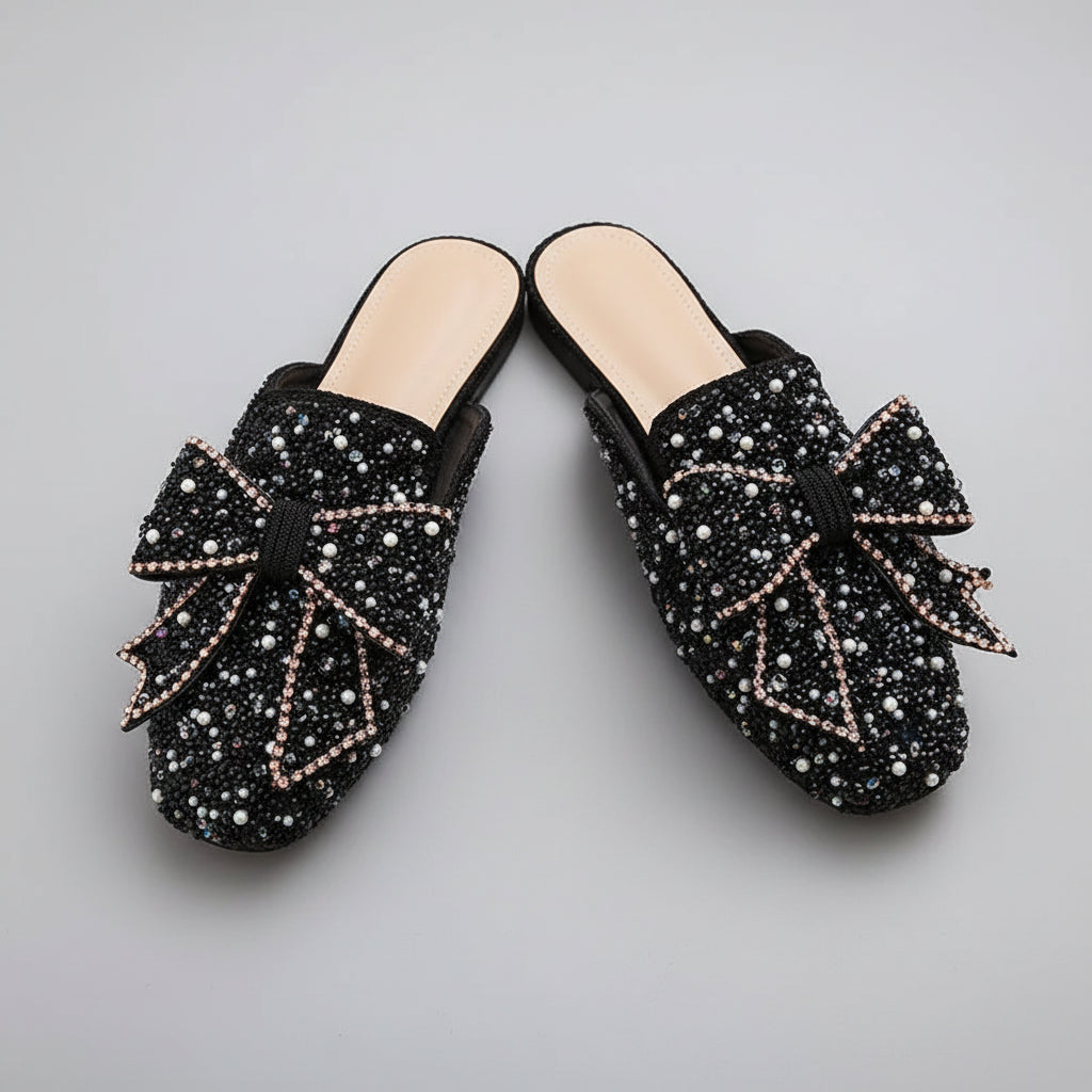 A pair of women's black mule loafers covered in rhinestones and bow details, resting inside a white shoebox.